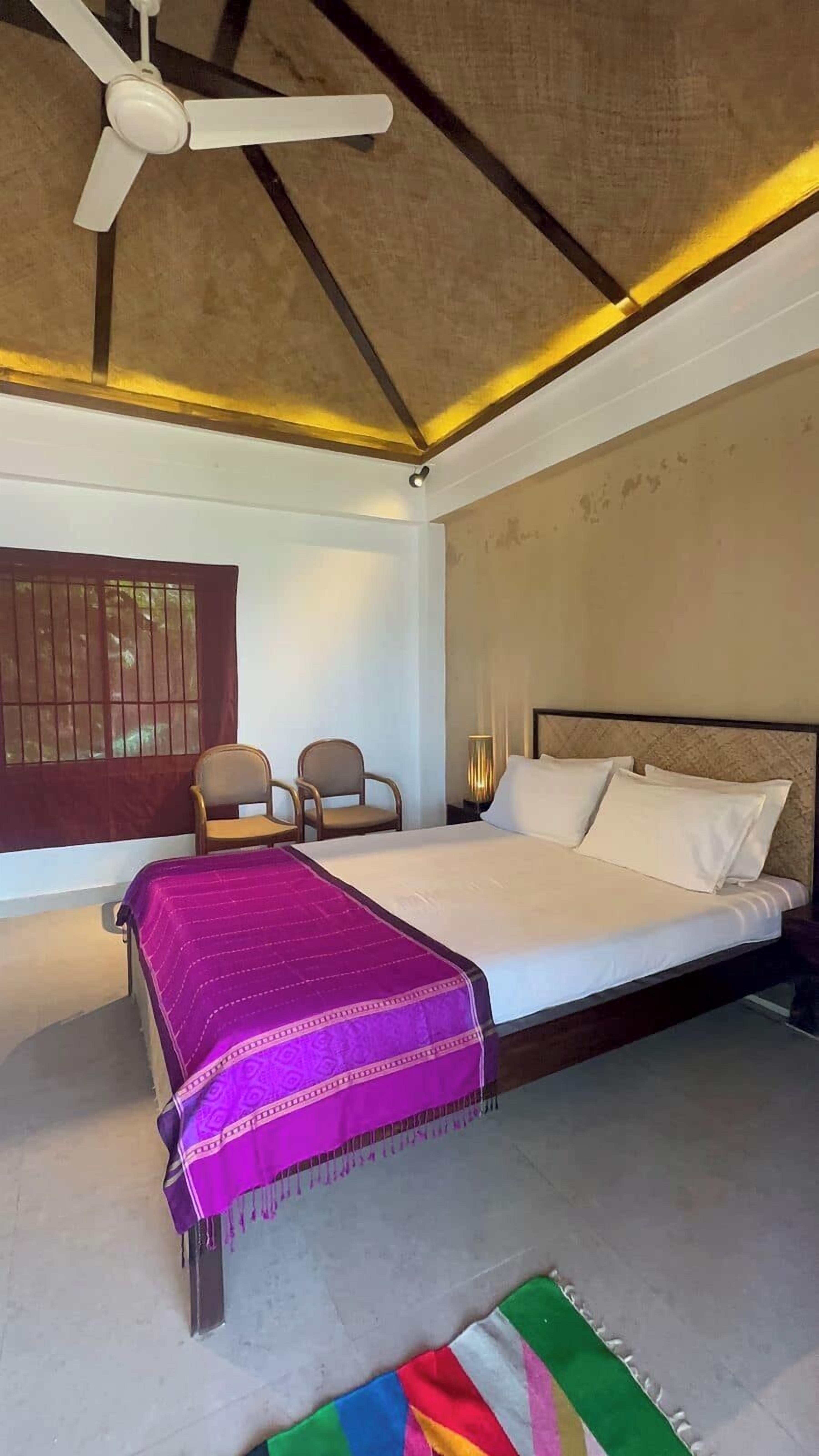 Un dormitorio con cama, una silla, un ventilador de techo y una ventana con barrotes.