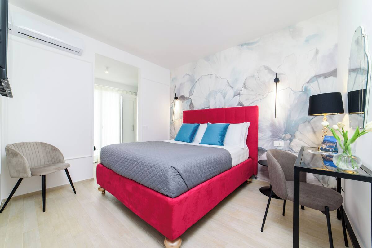Quarto moderno com cama vermelha, almofadas cinzentas e azuis, uma escrivaninha com luminária e um vaso com flores.