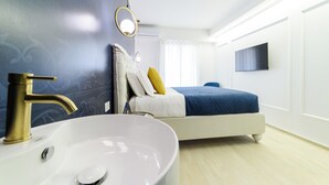 A modern hotel room with a large white sink, a gold faucet, a bed with a blue blanket, and a flat-screen TV mounted on the wall. - Shamika Luxury Rooms (Montecorvino Pugliano)