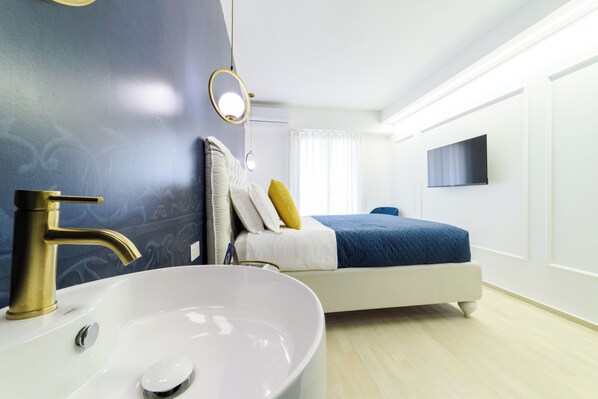 Una habitación de hotel moderna con un lavamanos blanco grande, un grifo dorado, una cama con una colcha azul y un televisor de pantalla plana montado en la pared.