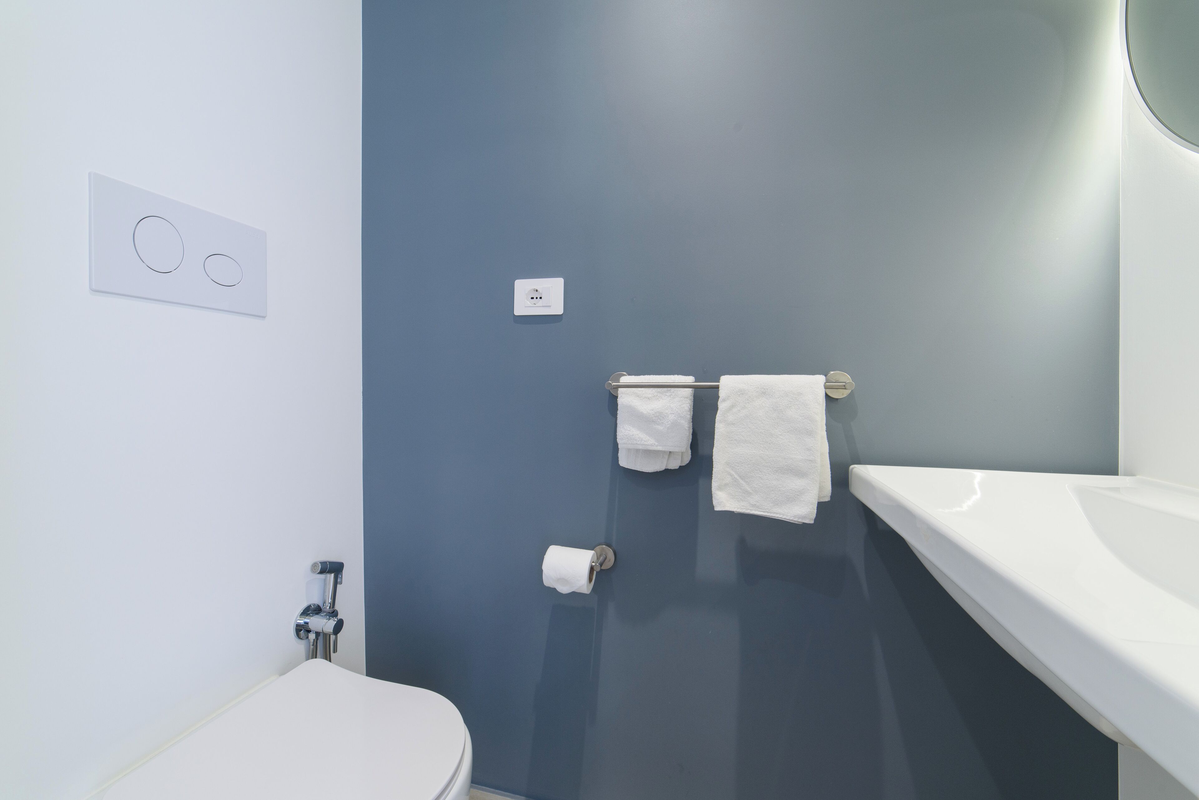 Ein Badezimmer mit einer weißen Toilette, einem Handtuchhalter mit zwei Handtüchern, einem Toilettenpapierhalter und einem an der Wand montierten Lichtschalter.