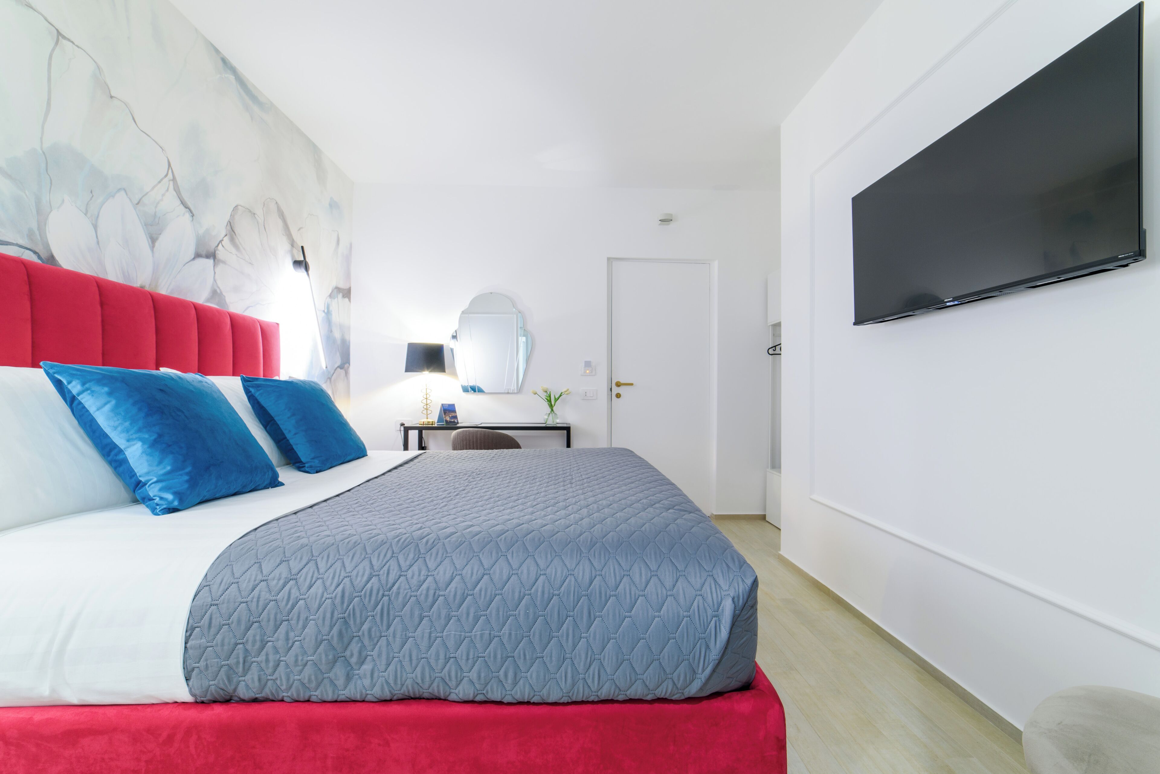 A modern hotel room with a large bed, a flat-screen TV, a desk, and a mirror.