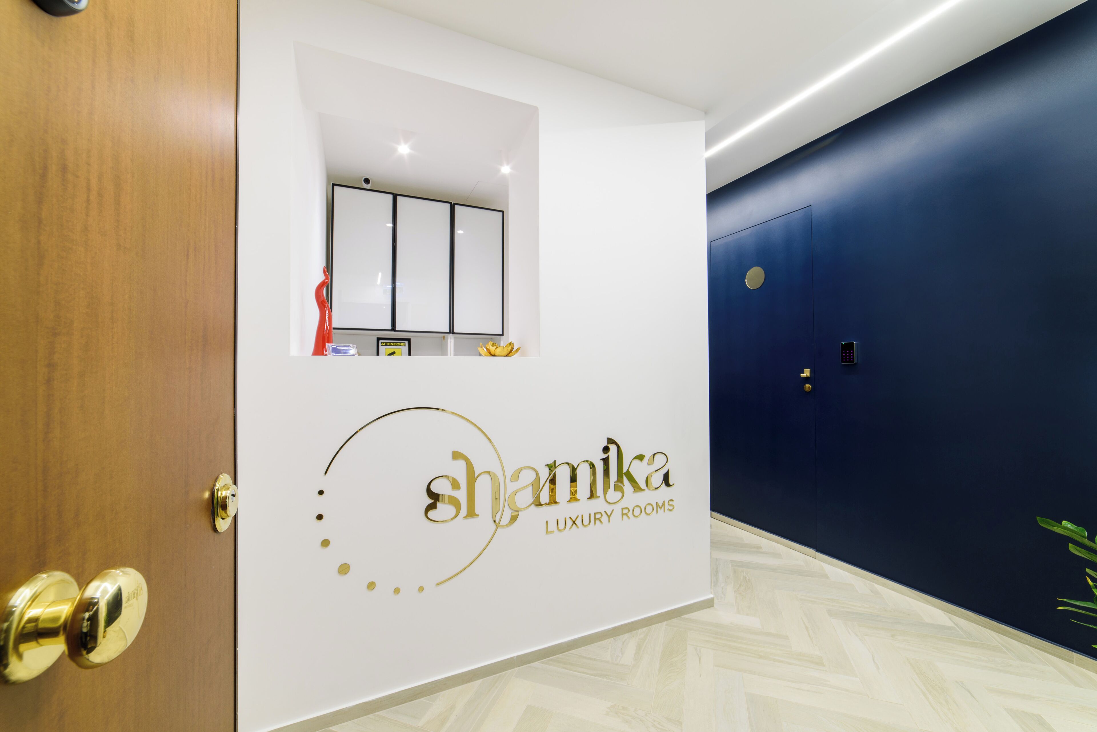 A hotel reception area with a white wall featuring the logo "shamika LUXURY ROOMS" and a door with a golden handle.