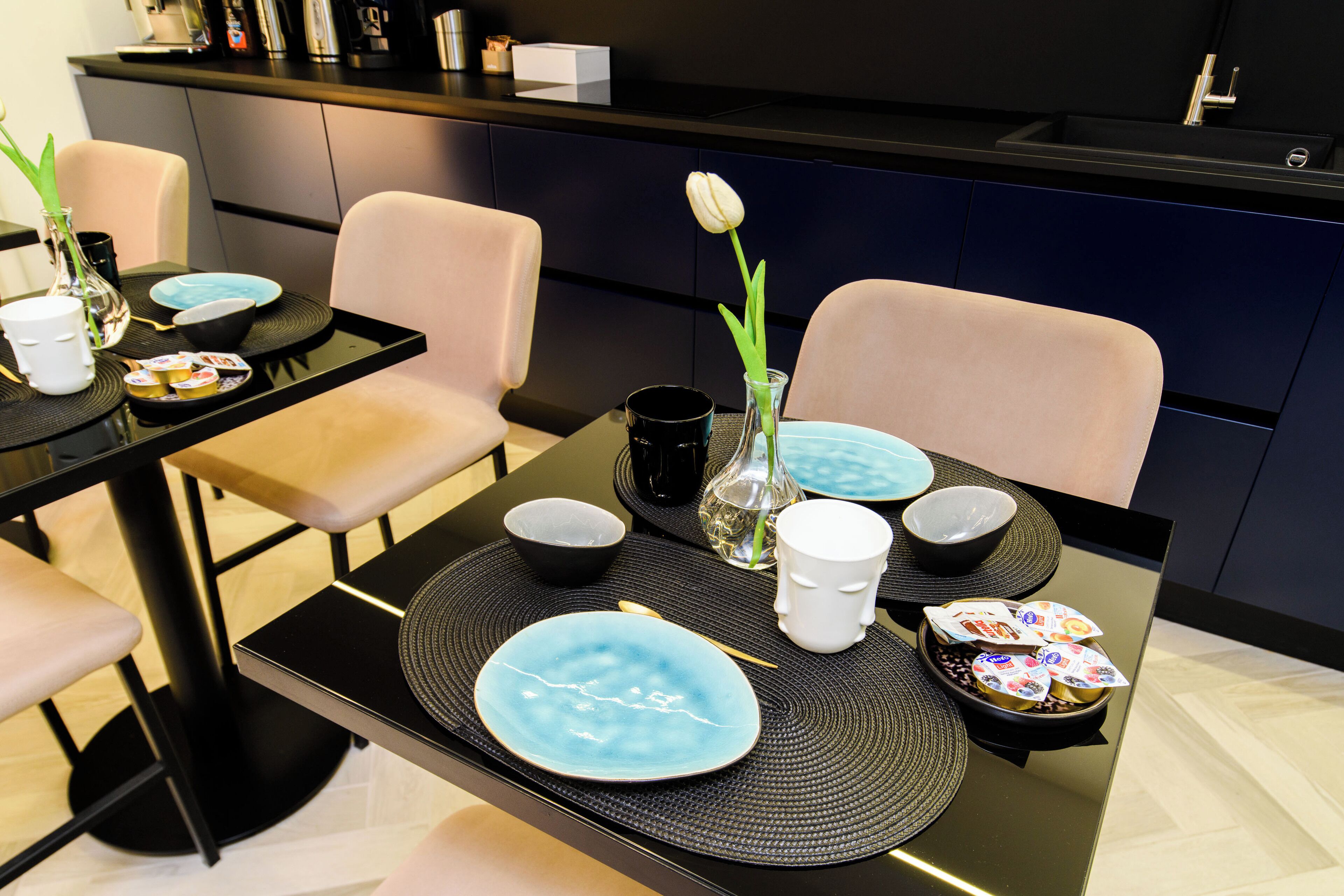 A modern dining area with two tables set for a meal, featuring blue plates, bowls, and a decorative vase with a flower.