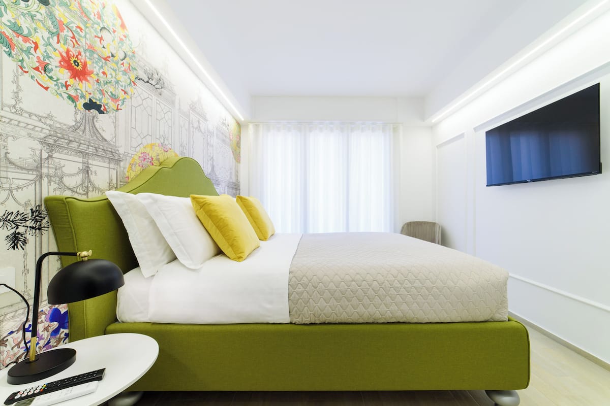 Quarto bem arrumado com uma cama verde, roupa de cama branca e uma televisão fixada na parede.