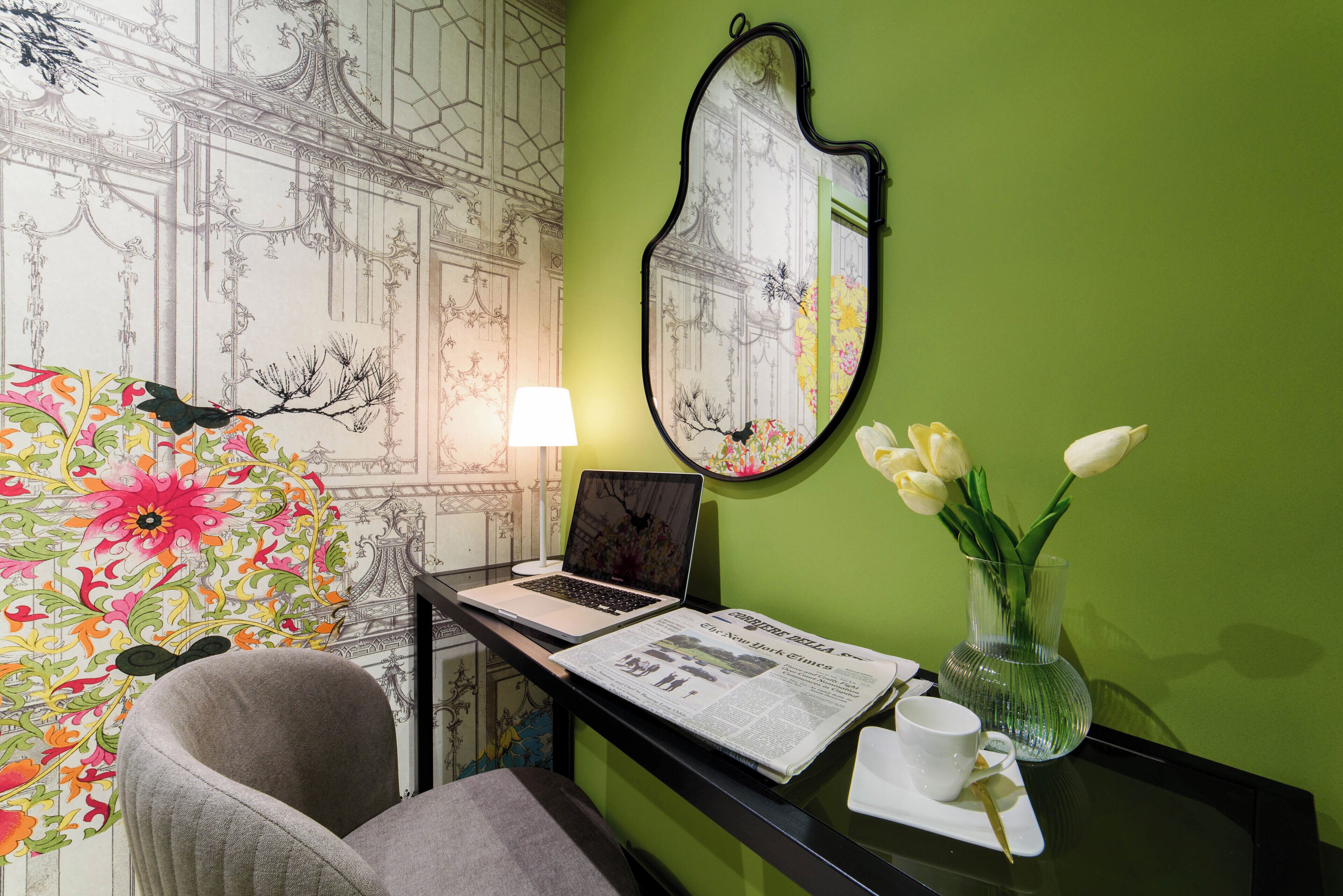 A room with a green wall, a mirror, a laptop, a newspaper, a vase with flowers, and a chair.