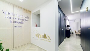 A hallway with a white wall featuring a logo and text, a herringbone-patterned floor, and a door leading to a room. - Shamika Luxury Rooms (Montecorvino Pugliano)