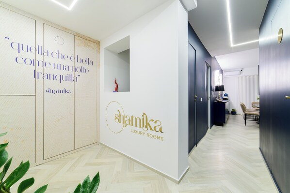 A hallway with a white wall featuring a logo and text, a herringbone-patterned floor, and a door leading to a room. - Shamika Luxury Rooms (Montecorvino Pugliano)