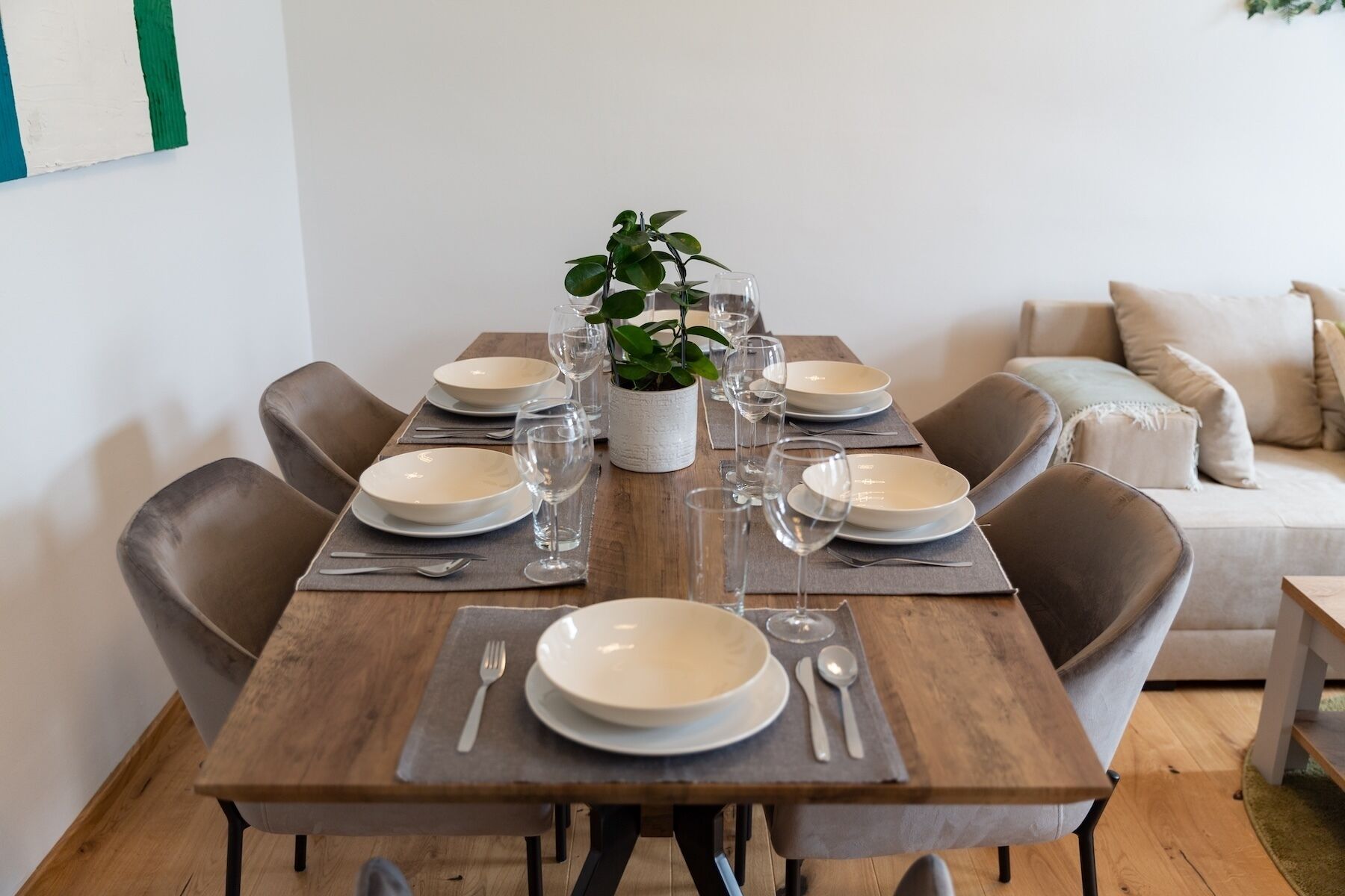 A dining table set with plates, glasses, and cutlery, surrounded by chairs, with a plant centerpiece and a sofa in the background.