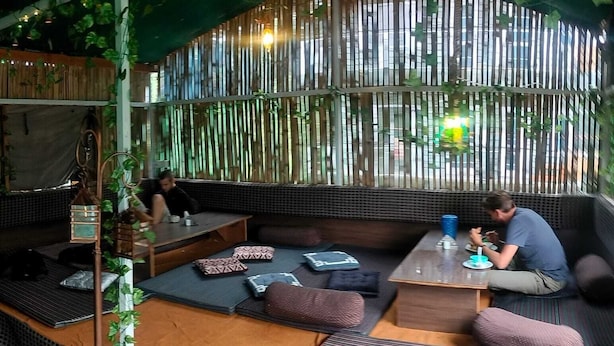 Un espacio al aire libre acogedor con bancos acolchados y una mesa baja, rodeado de follaje y paredes de bambú.