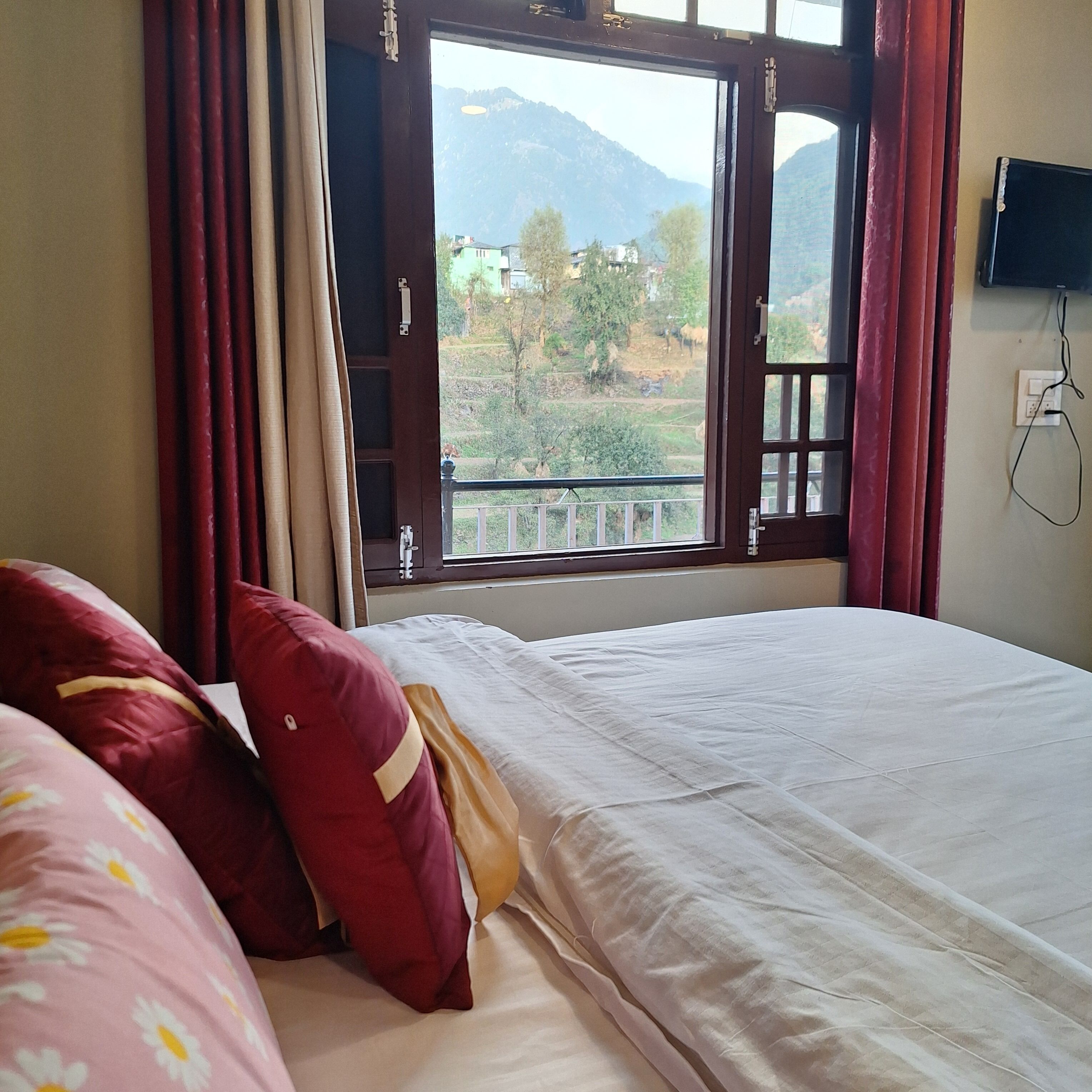 A bedroom with a bed, red and beige pillows, a window with a view of greenery and mountains, and a TV mounted on the wall.
