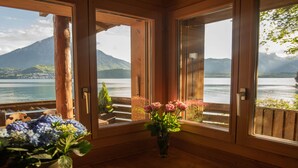 Una stanza in legno con pavimento in legno, un tavolo anch'esso in legno su cui si trova una pagnotta e un coltello, un vaso con dei fiori, e vista su un lago e montagne attraverso porte aperte.