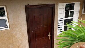 A brown wooden door with a metal handle, a window with white shutters, and a green plant beside the door. - Studio bungalow WiFi/ Hot Water in flourishing Drax Hall (Saint Ann's Bay)