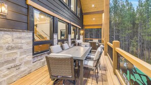 A wooden deck with a dining table and chairs, large windows, and a view of a forest. - Midnight Pines Lodge+HotTub+Cent AC+20mYellowstone (Island Park)