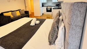A modern bedroom with a large bed, a grey headboard, and a view into the kitchen. - Stay Near Heathrow! Pristine, Convenient & Only 5 Mins to the Terminals (Staines)
