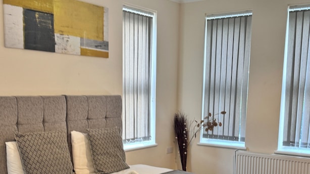A modern bedroom with a large bed, grey and white bedding, a grey headboard, and a wooden floor. - Stay Near Heathrow! Pristine, Convenient & Only 5 Mins to the Terminals (Staines)