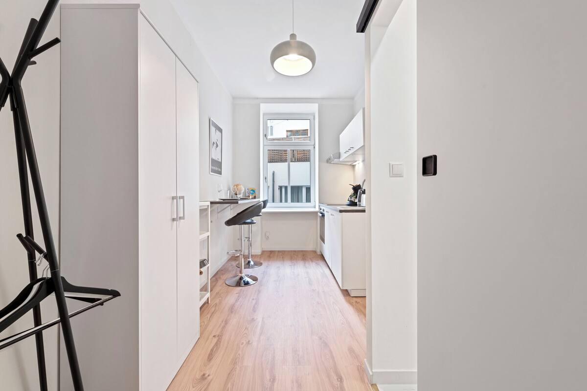 A narrow hallway with a wooden floor, white walls, and a window at the end leading to a kitchen area.