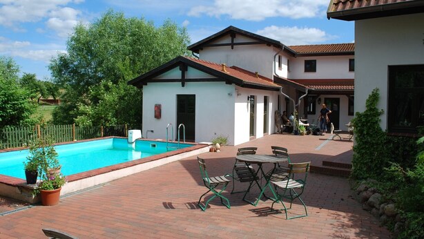 Una piscina con zona de patio, mobiliario exterior y un pequeño edificio con puerta.