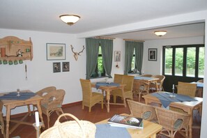Een eetruimte met rieten meubels, groene gordijnen en ingelijste foto’s aan de muren.