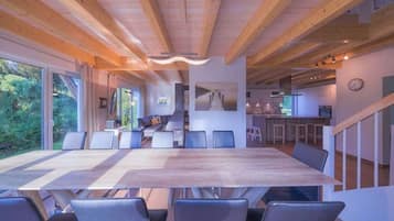 A spacious dining area with a long wooden table and modern chairs, a kitchen in the background, and large windows offering a view of the outdoors.