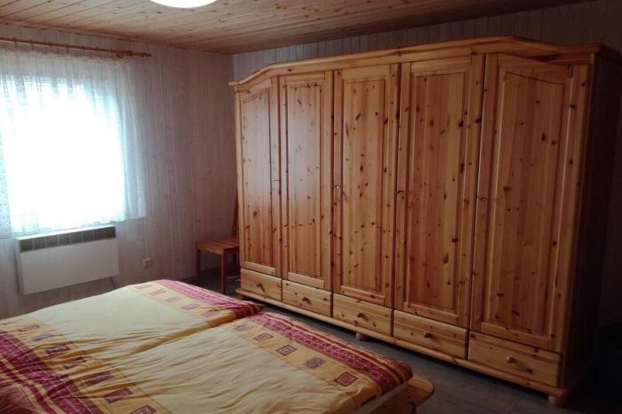 Quarto com guarda-roupa de madeira, cama com colcha estampada e janela com cortinas.