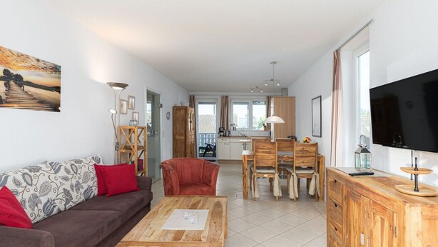 A modern living room with a sofa, a coffee table, and a TV. There is a dining area with a table and chairs. The room has a large window and a kitchen in the background. - Appartement im Ferienpark in Rechlin (Rechlin)