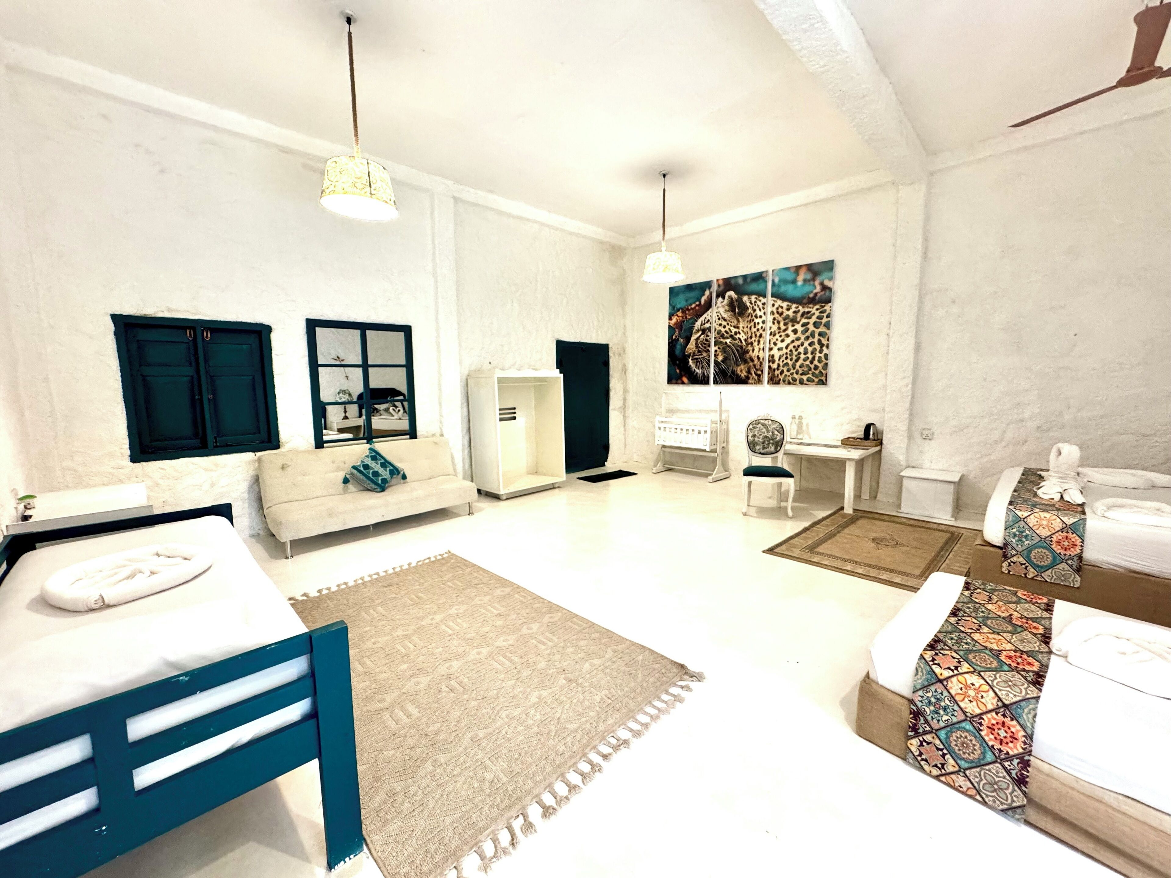 A spacious room with a bed, a sofa, a desk, and a chair. There are two windows with dark frames, a ceiling fan, and a painting on the wall.