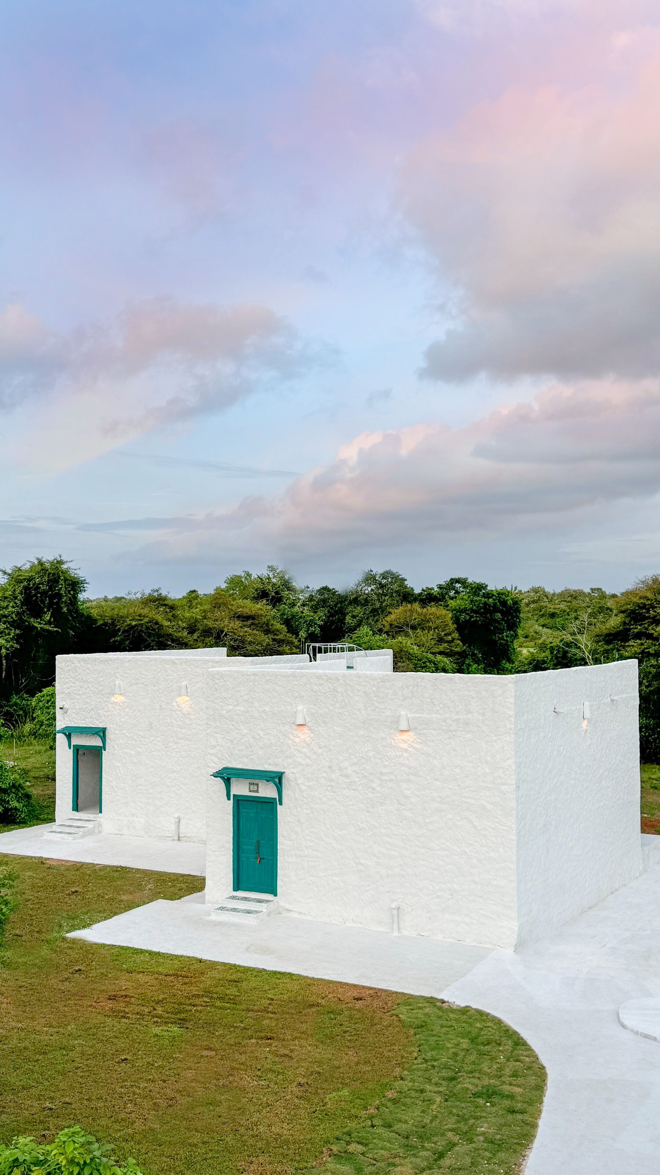 A white building with a green door and windows, surrounded by greenery and a cloudy sky.