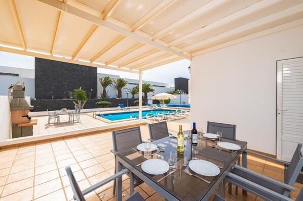 A covered outdoor dining area with a pool in the background.