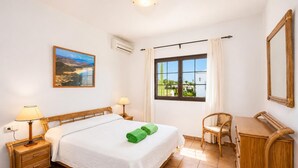 Ein Hotelzimmer mit einem Bett, zwei Nachttischen, einem Schreibtisch, einem Sessel, einem Fenster mit Vorhängen, einer Klimaanlage und einem Bild an der Wand.