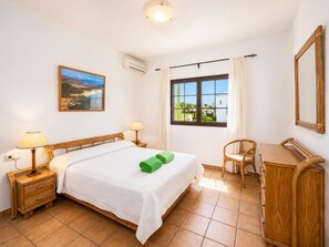 Une chambre d’hôtel comprenant un lit, deux tables de chevet, un bureau, une chaise, une fenêtre avec des rideaux, un climatiseur et un tableau au mur.