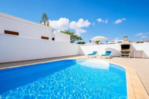 Une piscine avec des chaises longues, un espace barbecue et un mur blanc agrémenté de fenêtres.