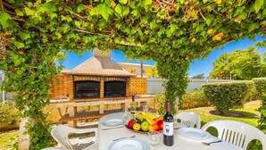 Un área de comedor al aire libre con un horno de ladrillo, sillas de plástico blanco y una mesa dispuesta con platos, cubiertos y una botella de vino.