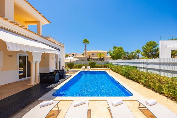 Piscina rectangular con sillas de descanso y sombrillas, rodeada por una cerca blanca y follaje.