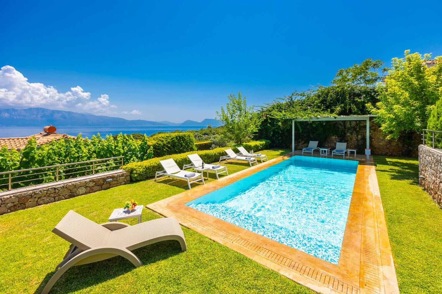 Una piscina rectangular con agua azul cristalina, rodeada por un muro de piedra y un área verde.
