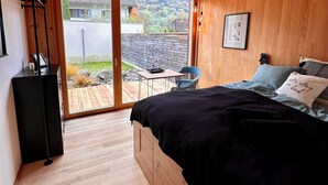 A bedroom with a wooden ceiling, a bed with a black bedspread, a desk with a chair, and a view of a garden and buildings outside.