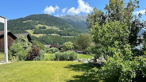 A grassy yard with a wooden cabin, a tree, and a mountain range in the background.