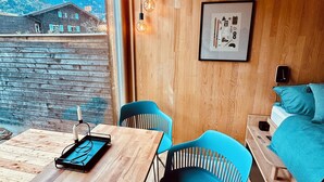 A wooden cabin interior with a wooden table, two blue chairs, a bed with teal bedding, and a framed picture on the wall.