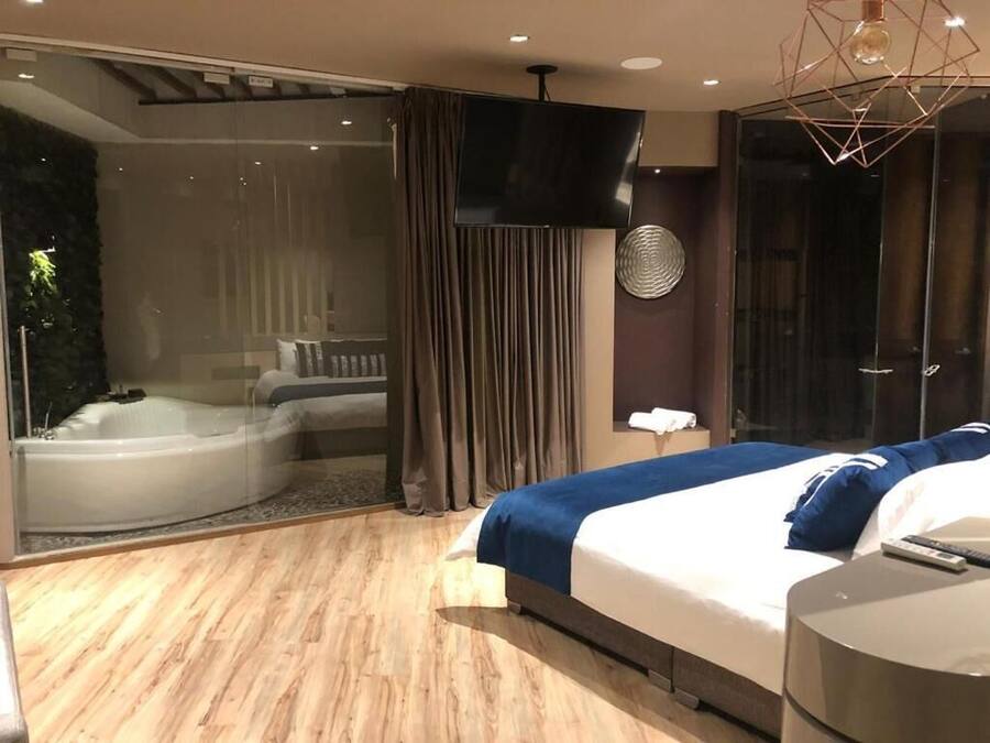Quarto de hotel moderno com uma cama grande, hidromassagem em vidro e uma televisão de tela plana.