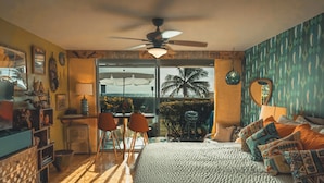 Une chambre avec un lit, un ventilateur de plafond, un bureau avec des chaises et une vue sur les palmiers, visible à travers une porte vitrée.