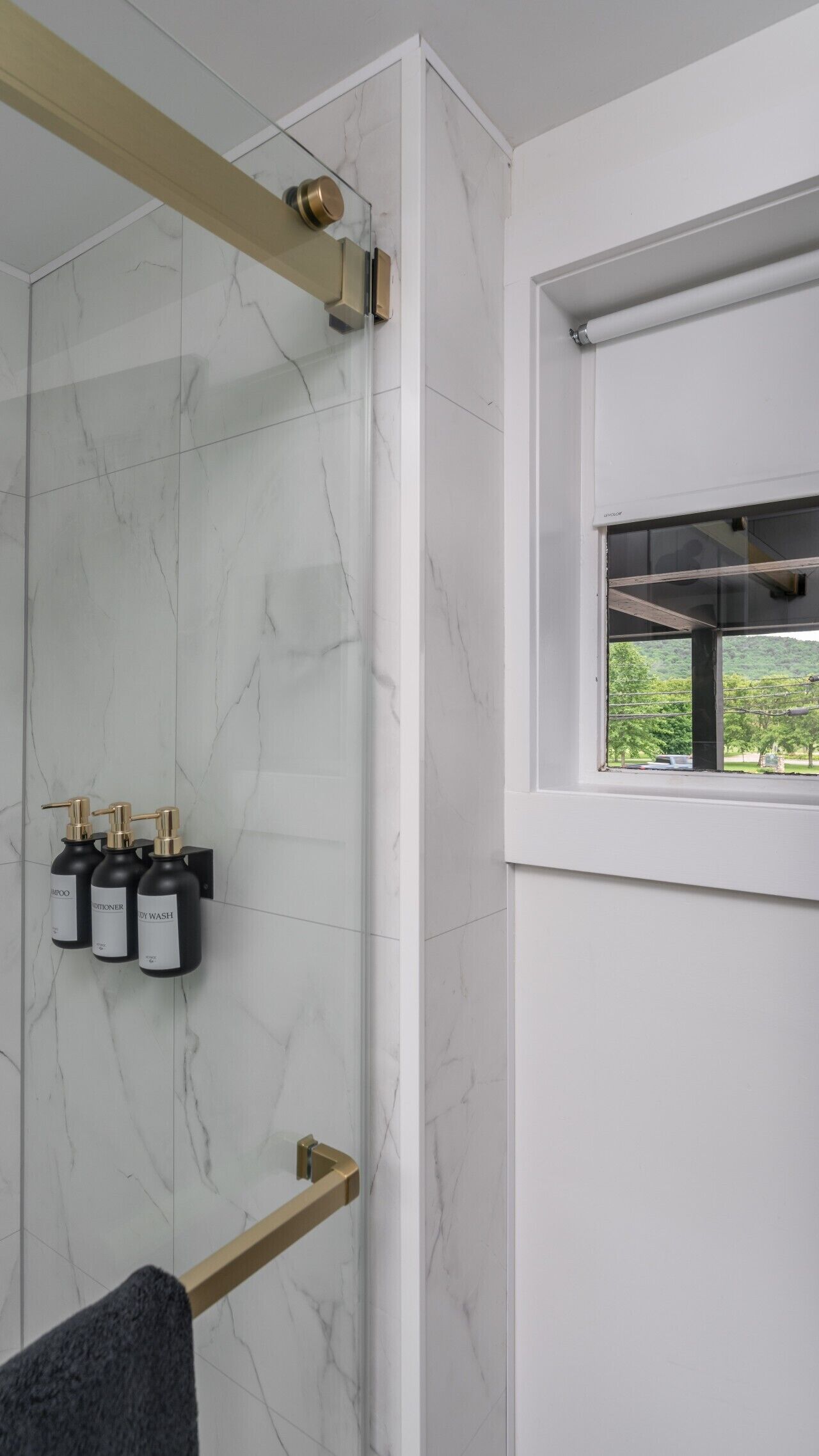 A modern bathroom with a marble wall, a glass shower enclosure, and a window with a view of greenery.