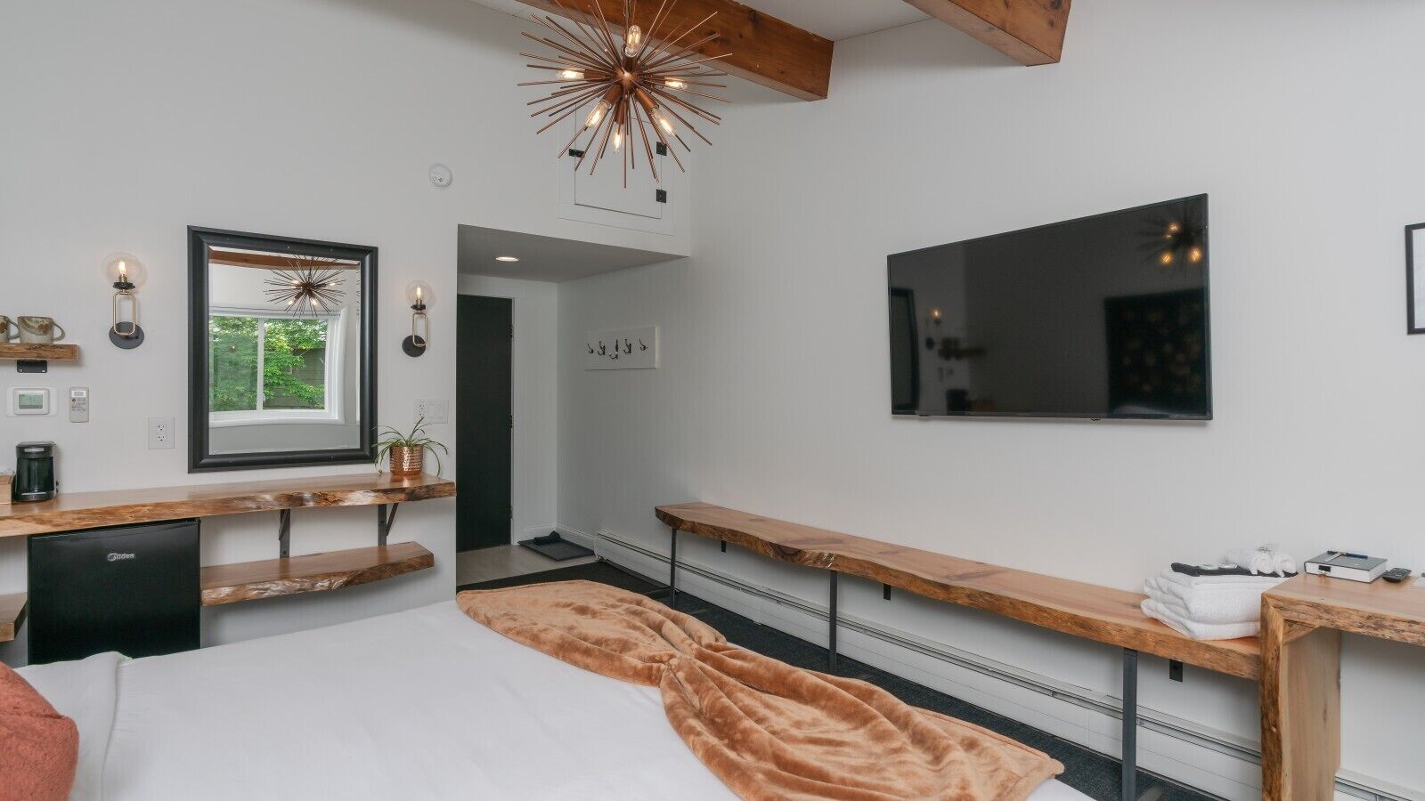 Een moderne slaapkamer met een bed, een houten bankje, een spiegel, een televisie en een plafondlamp.