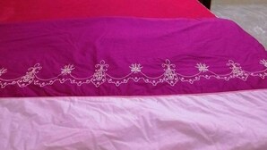 Ein ordentlich bezogenes Bett mit einer roten und violetten Tagesdecke, einem hölzernen Kopfteil und einem kleinen Nachttisch daneben.