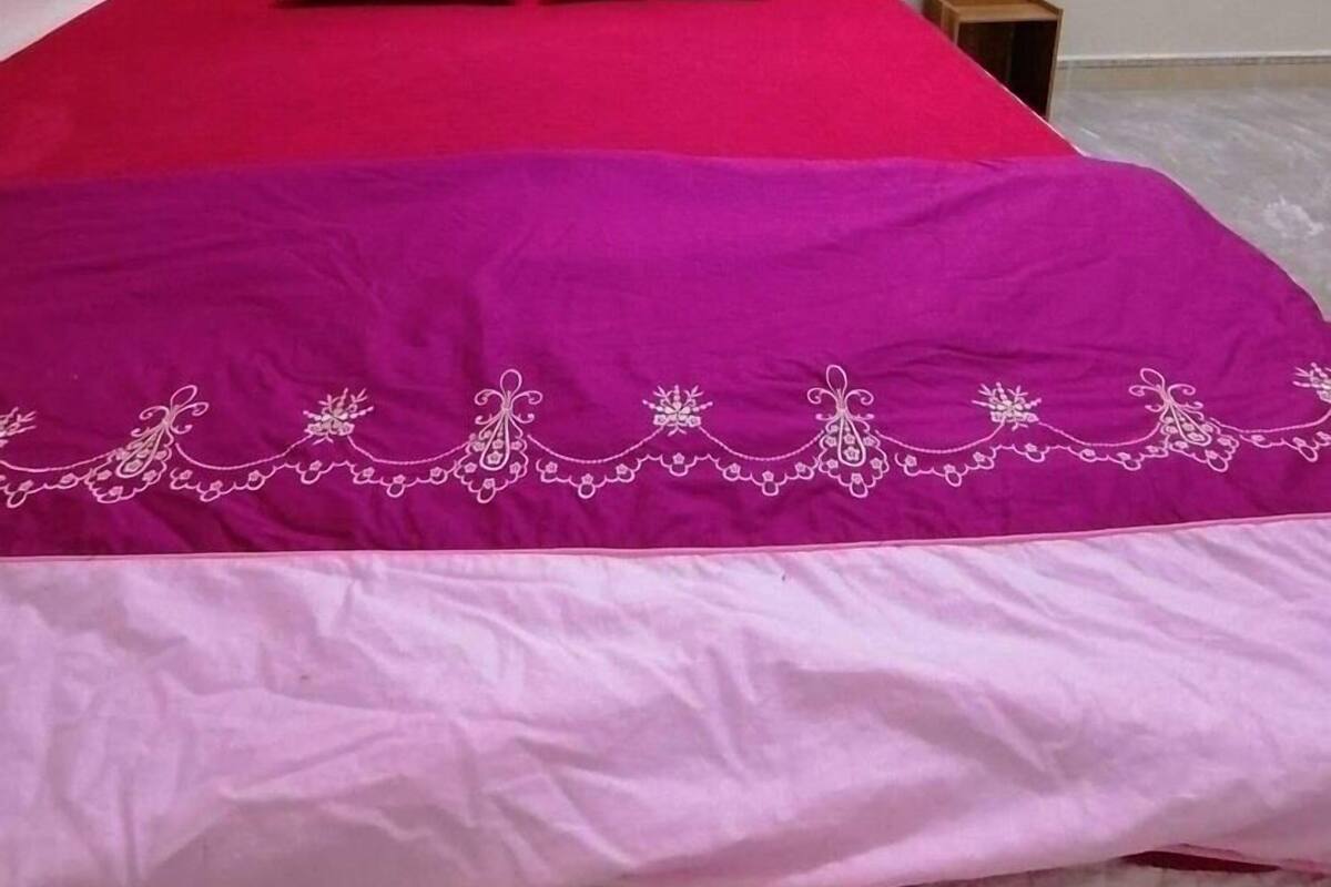 A neatly made bed with a red and purple bedspread, a wooden headboard, and a small wooden nightstand beside it.