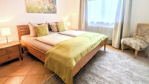 Un dormitorio con cama, mesitas de noche, lámpara, un cuadro, una ventana con cortinas y una silla.
