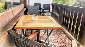 Una mesa de madera con un recipiente de vidrio en un balcón con sillas de mimbre y una barandilla metálica.