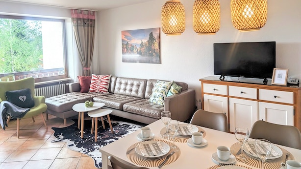 Una sala de estar moderna con un sofá, una mesa de comedor preparada para dos personas y un televisor de pantalla plana.