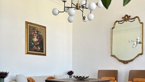 Een woonkamer met een ronde houten tafel, twee fauteuils, een bank, een kroonluchter, een grote spiegel en een bloemschilderij aan de muur.