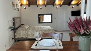 Een eettafel gedekt met borden en wijnglazen, een houten plafond, een bed met een geborduurde spreidek en een potplant met roze bloemen.