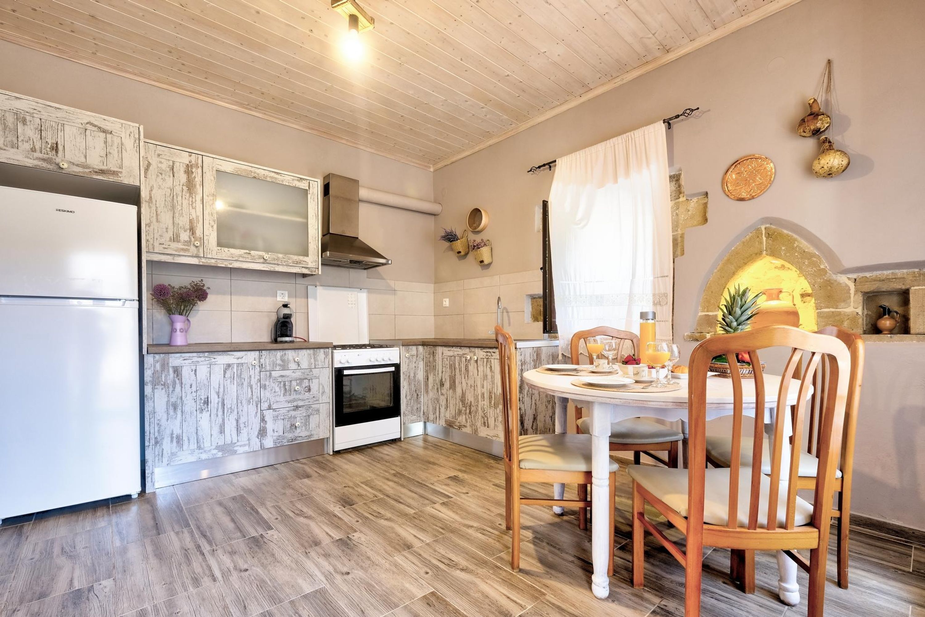 Een keuken met houten kastjes, een witte koelkast, een fornuis en een eettafel met stoelen.